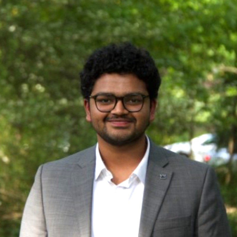 Portrait of Headshot of a young man in a grey blazer outside with foliage in the background.