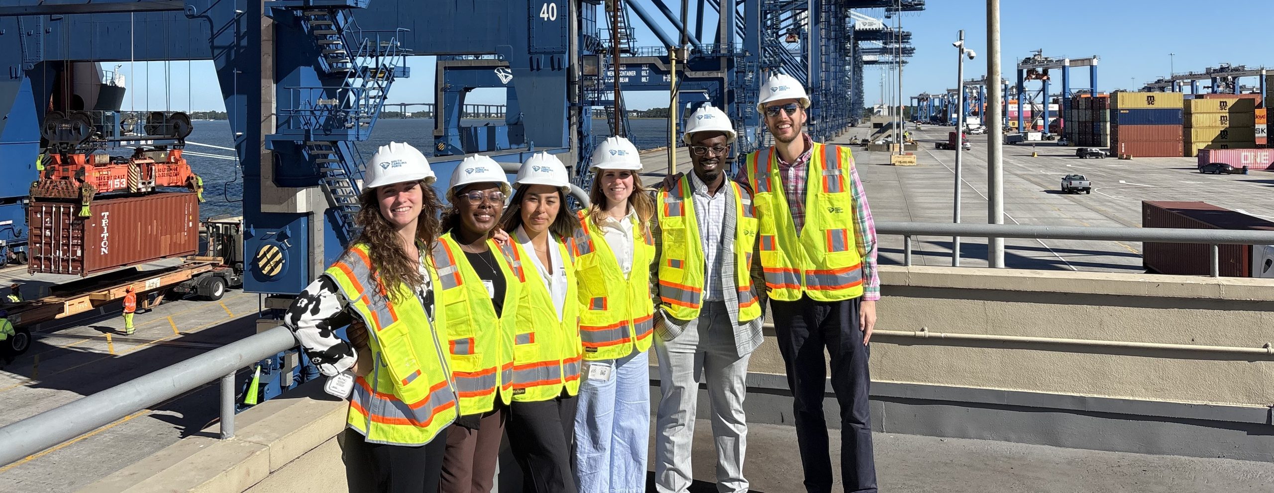 Group of six students wearing hard hats and yellow vests standing in front of industrial structures outside