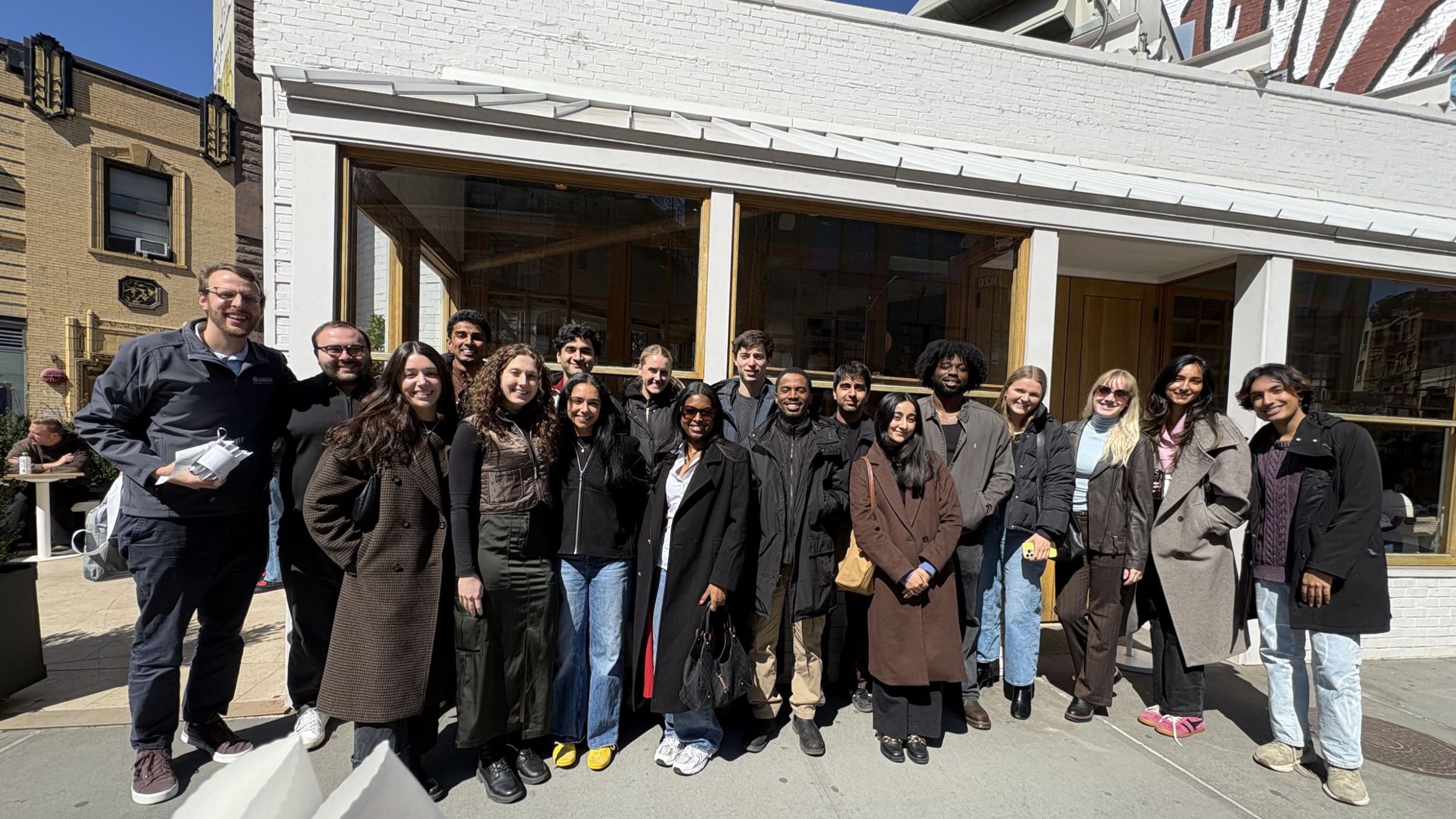 LHS Alumni Community Brunch group photo in NYC. Two rows of smiling people in coats standing on a sidewalk in the sunlight.