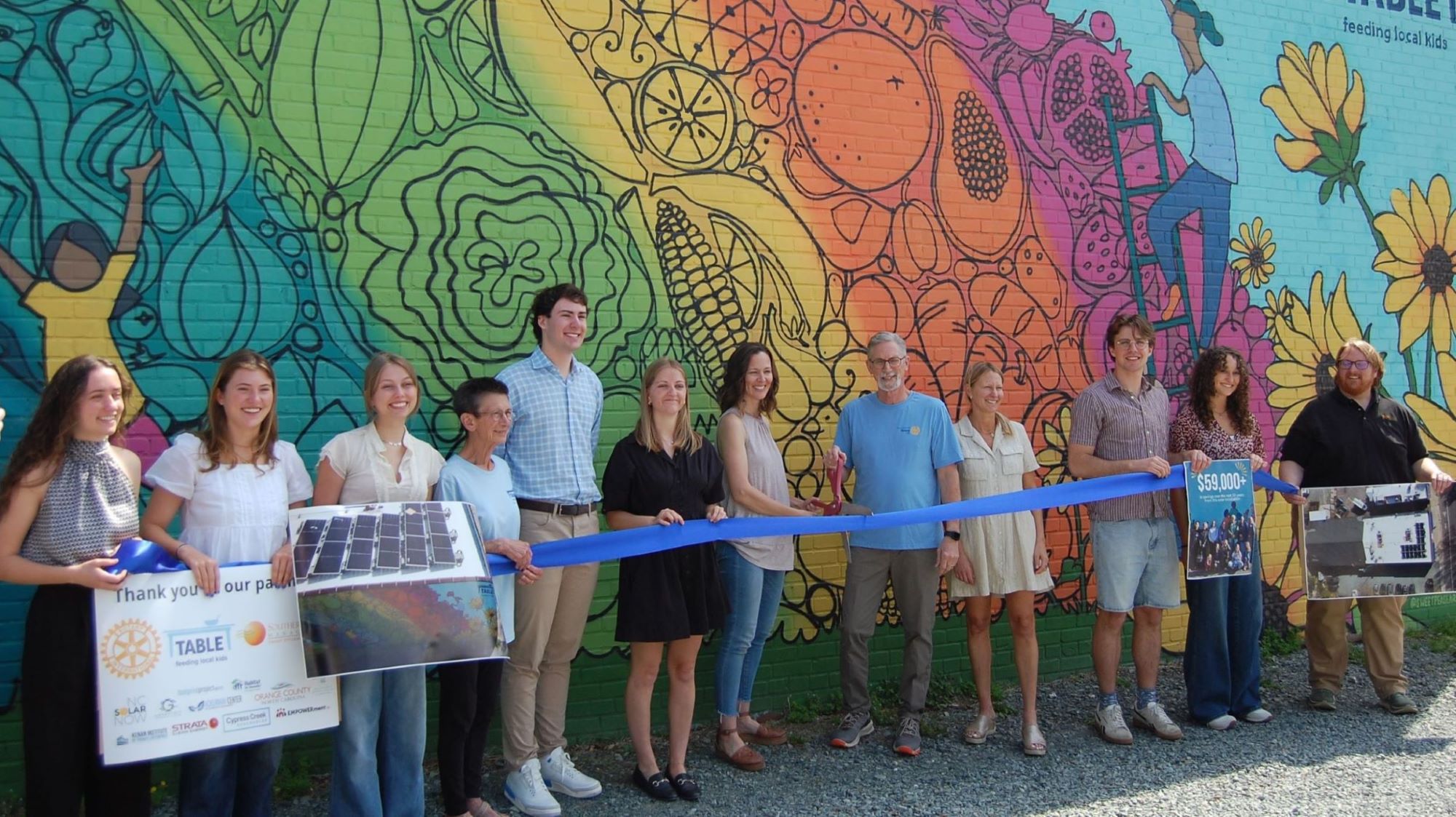 SolarEquity exec team and community partners smiling and standing in front of a colorful mural outside of a building, and holding a large blue ribbon to cut