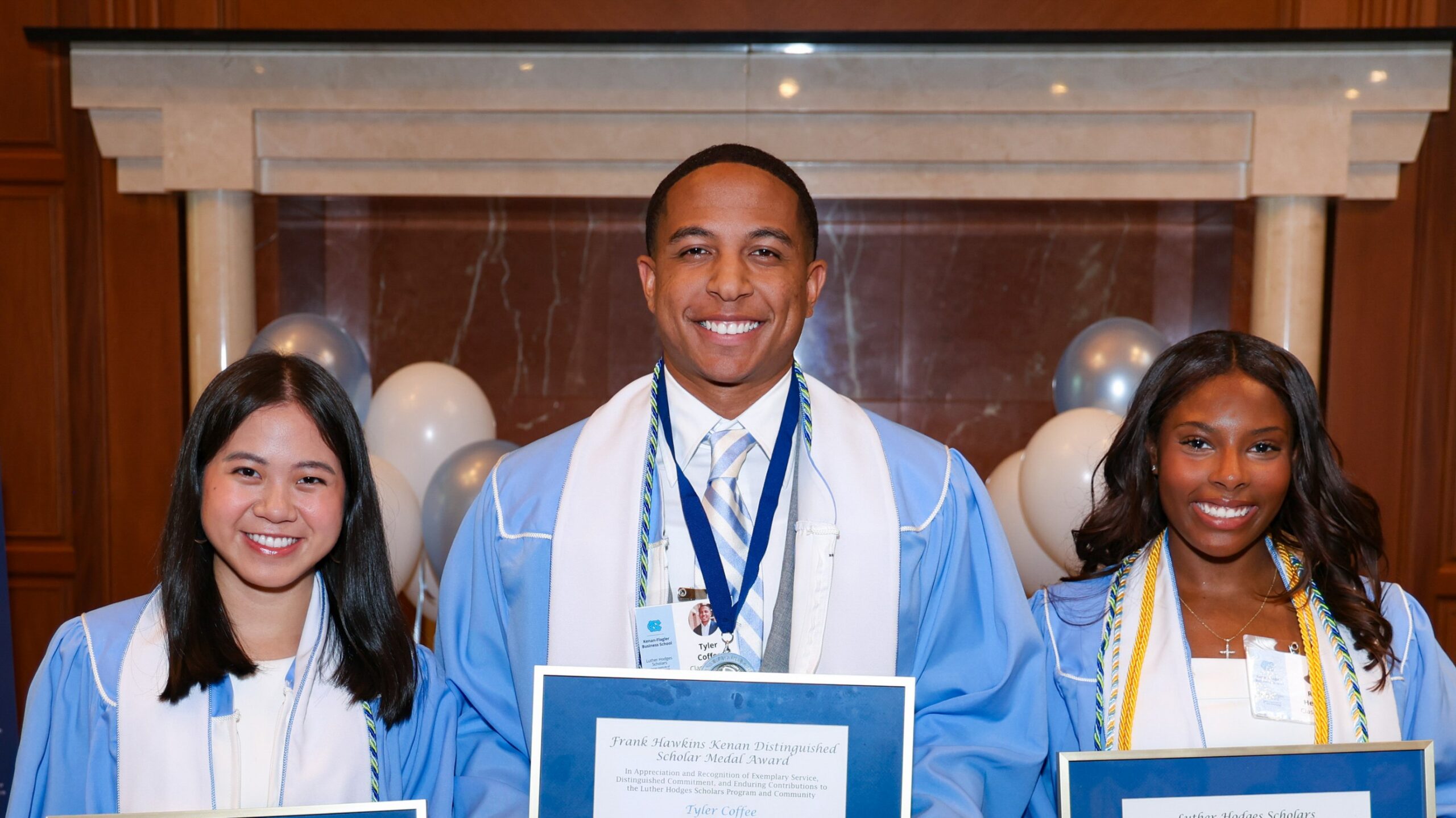 Three graduates in light blue gowns pose indoors holding framed certificates, with balloons and a celebration display in the background.