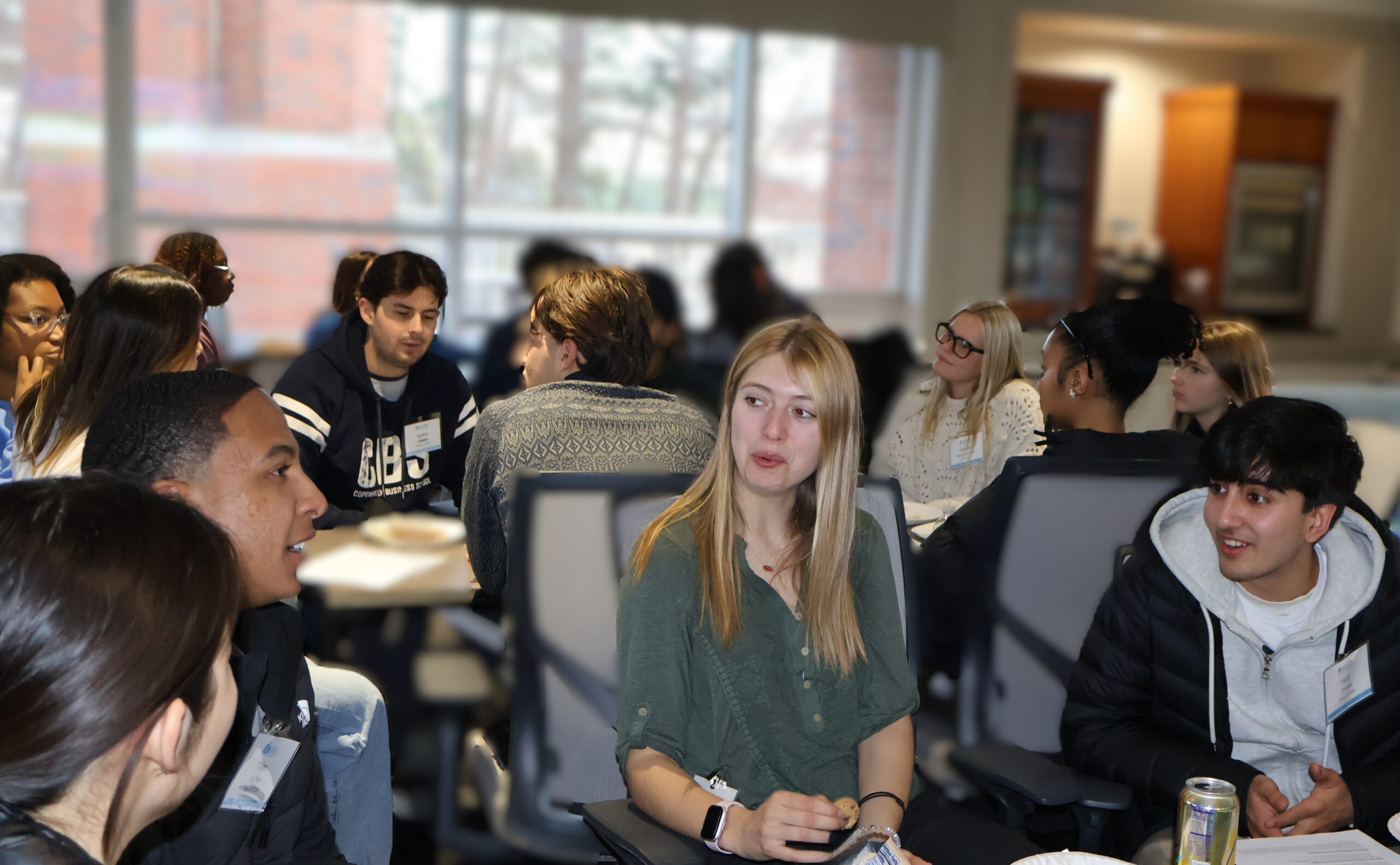 Hodges Scholars seated in small groups at tables, eating pizza and talking during an indoor Living Room Conversations event