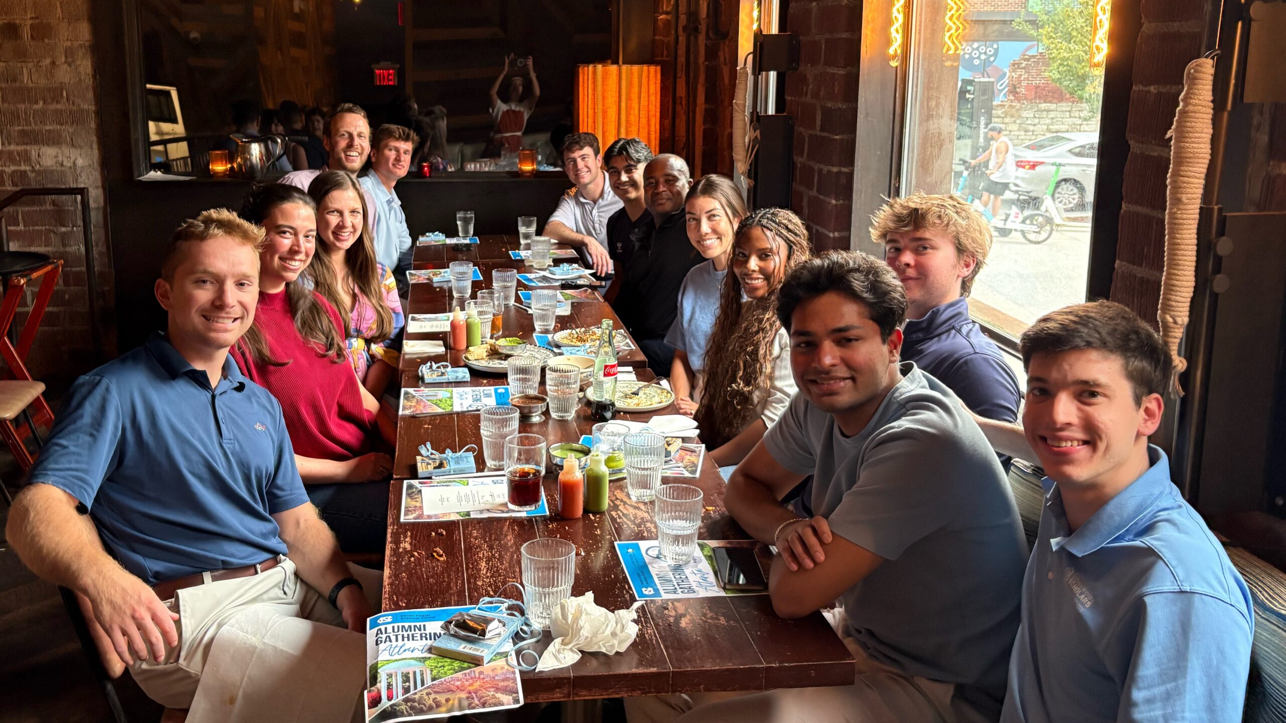 Luther Hodges Scholars Program alumni and supporters gather around a long table at a restaurant in Atlanta, Georgia, during an alumni networking dinner. Attendees from multiple graduating classes are seated together enjoying conversation and a shared meal, with event materials visible on the table and large windows overlooking the street.