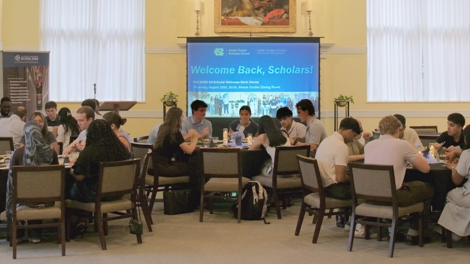 students seated at round tables in a bright banquet hall during a welcome-back dinner. A large presentation screen at the front of the room displays “Welcome Back, Scholars!” with Kenan-Flagler Business School and Luther Hodges Scholars Program branding.