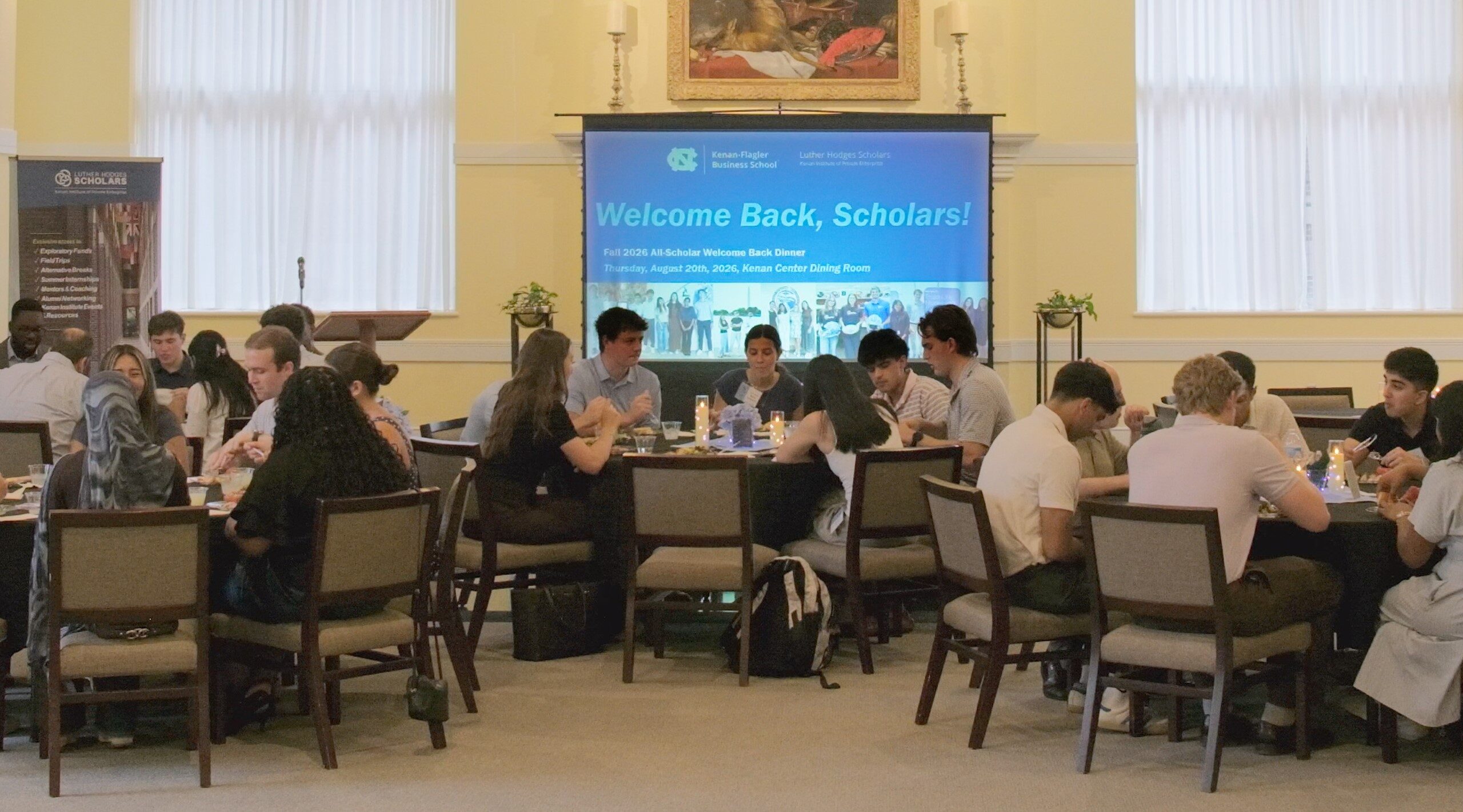 students seated at round tables in a bright banquet hall during a welcome-back dinner. A large presentation screen at the front of the room displays “Welcome Back, Scholars!” with Kenan-Flagler Business School and Luther Hodges Scholars Program branding.